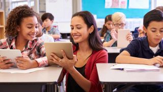 Smiling teacher kneels next to student's desk as they both look at tablet computer. 