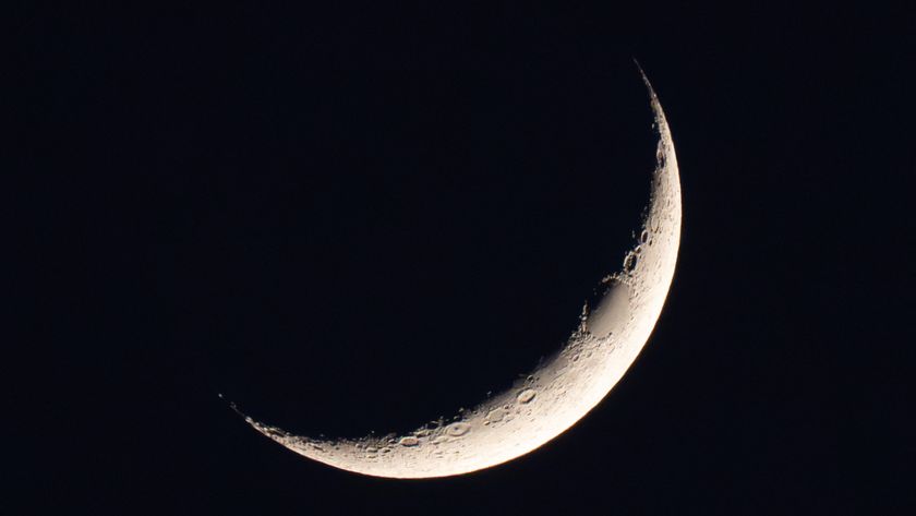 A photo of a thin waxing crescent moon shining in a black night sky. A dark oval is visible on the upper section of its sickle-like profile. Elsewhere, large craters can be seen pockmarking the illuminated wedge of the lunar disk.
