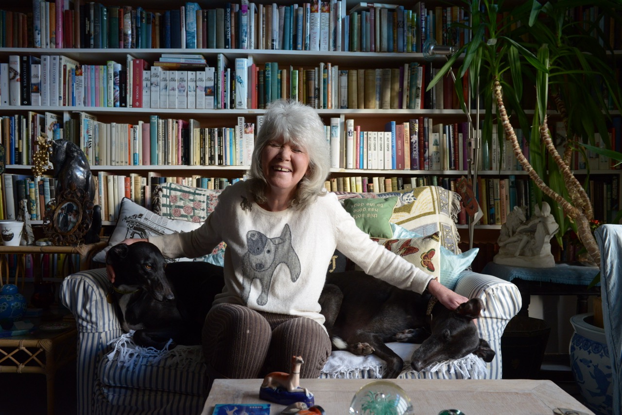 Jilly Cooper sits smiling on a striped sofa in front of a wall of bookshelves, flanked by two relaxed black greyhounds resting beside her. She wears a cream jumper with a greyhound illustration on it and is surrounded by cushions and books in a cosy, colourful living room.