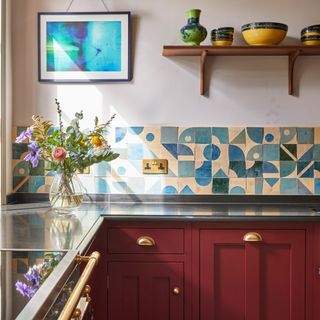 a kitchen countertop with dark red cabinets with brass cup pulls, and a striking blue graphic tiled splashback and a vase of flowers