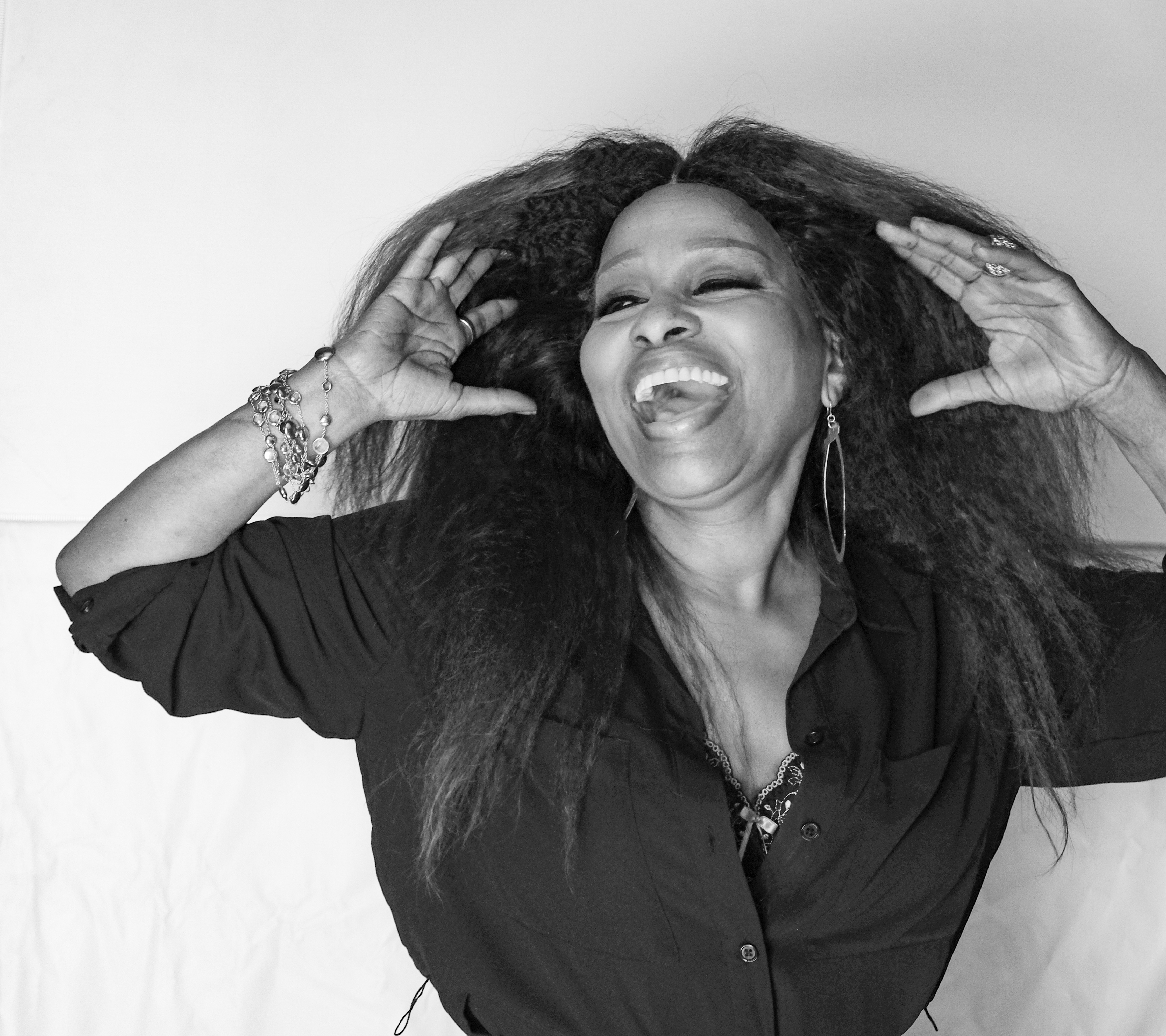 Black and white portrait of Chaka Khan laughing with her head thrown back, both hands raised to her voluminous hair.