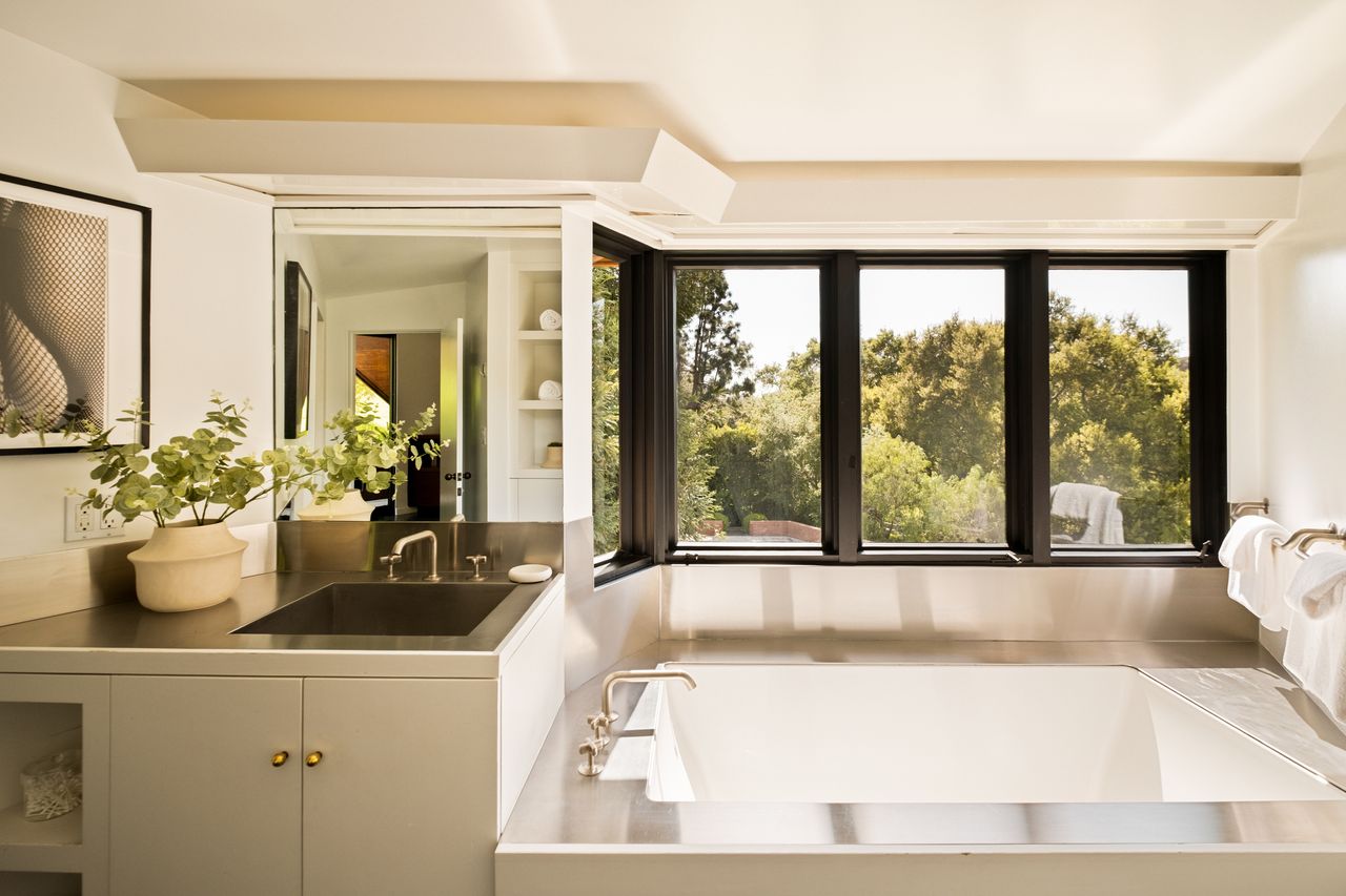 Image of a white bathroom showing a sink and vanity next to a tub. There are windows above the tub that are trimmed in black. The countertop and tub edge are stainless steel. A plant in a white vase is on the sink counter.