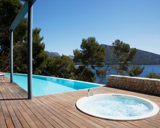 A pool and hot tub on a sunny, wooden terrace with a view of the sea, trees and hills in the background