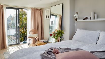 Pink drapes over sliding doors in bedroom with white bedding, full length mirror, rattan chair and greenery indoors