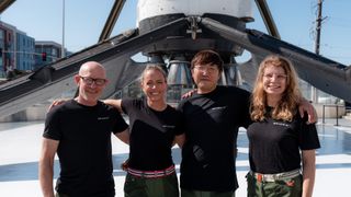 four smiling people in black t-shirts stand in front of a rocket 