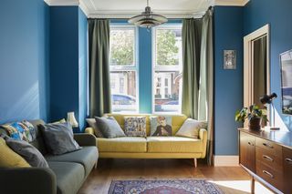 A deep teal living room with yellow and green sofas, a mid-century style sideboard and a rug