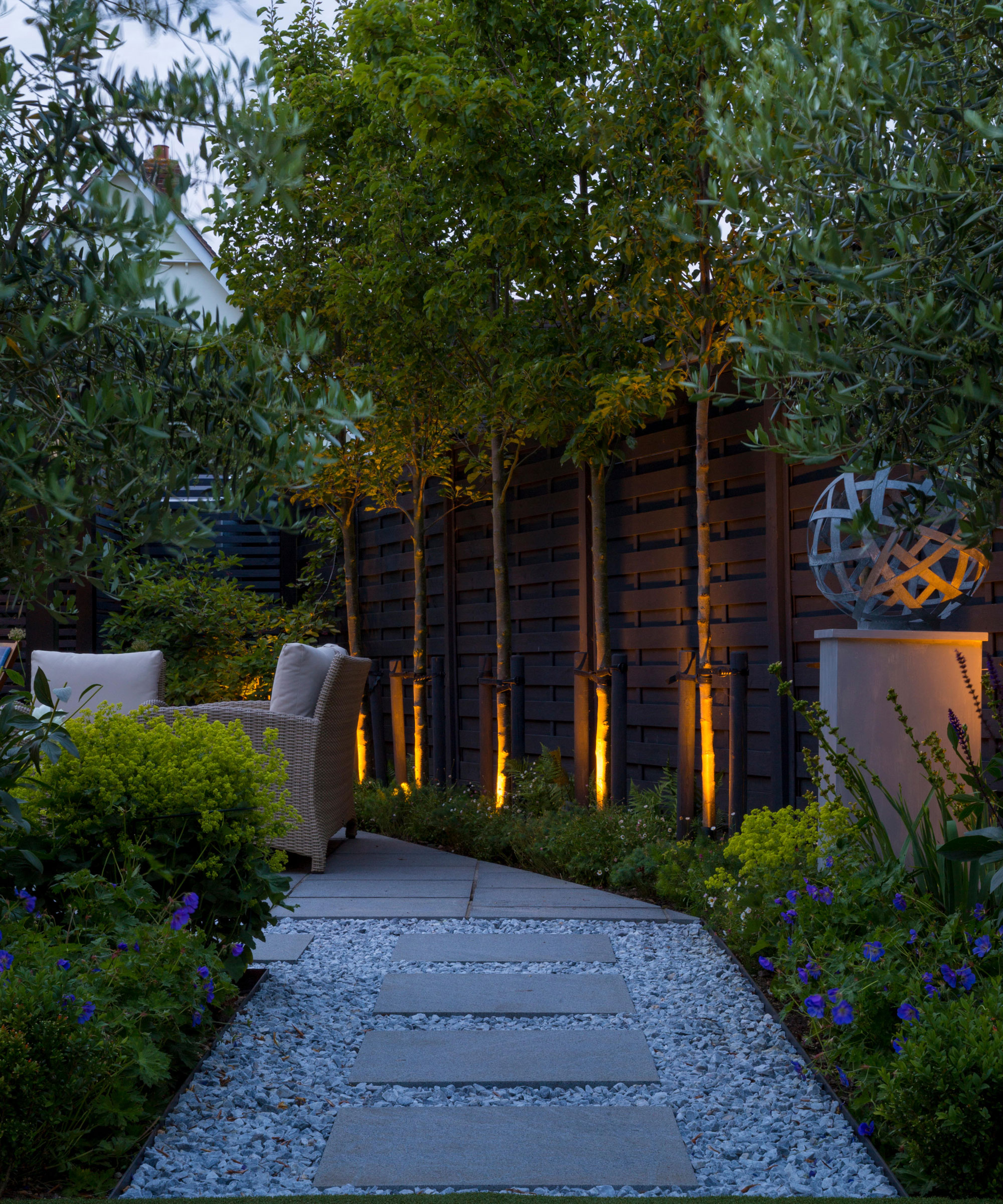 uplit pleached trees and uplit garden sculpture with garden chairs and gravel path with pavers