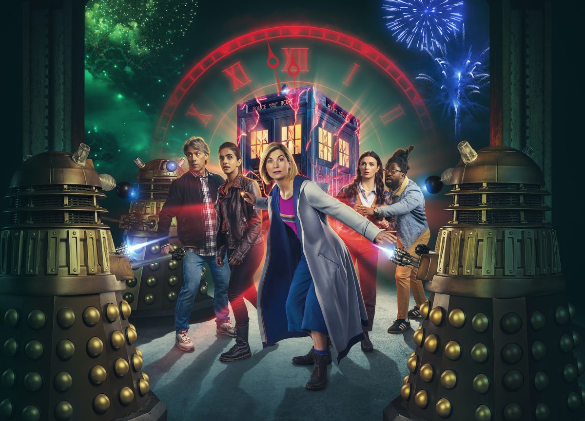 Jodie Whittaker as The Doctor in a preview image for Eve Of The Daleks featuring Dan (John Bishop), Yaz (Mandip Gill), Sarah (Aisling Bea) and Nick (Adjani Salmon) , standing in front of the Tardis and surrounded by Daleks, while a clockface in the background ticks towards midnight and fireworks erupt in the sky