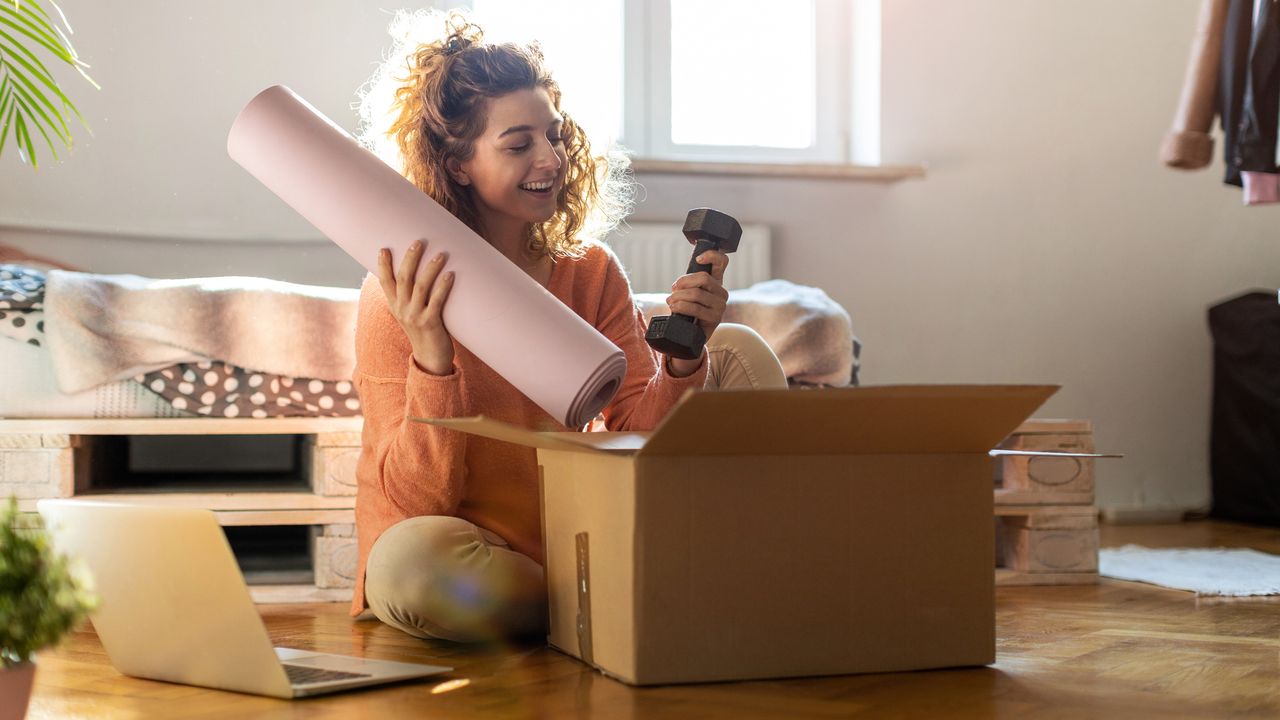 Woman sits cross-legged on floor in front of carboard box holding a rolled up yoga mat in one hand and a light dumbbell in the other