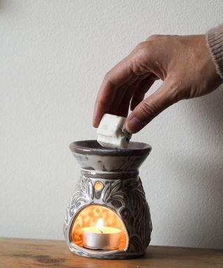 Someone placing a solid wax melt into an ornate white and gray wax melter heated by a lit tealight which is casting a glow inside the item