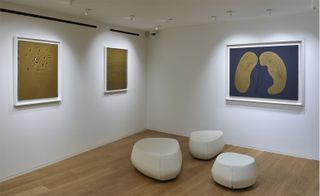 Inside the exhibition with three canvas paintings on a wall and three stone-style seats.
