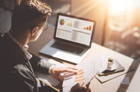 Young finance market analyst in eyeglasses working at sunny office on laptop while sitting at wooden table.Businessman analyze document in his hands.Graphs and diagramm on notebook screen.Blu