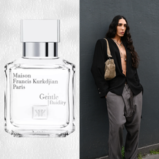 A woman wearing a black button up with a deep v neck and gray jeans leaning against a black wall. She is next to a bottle of unisex perfume.