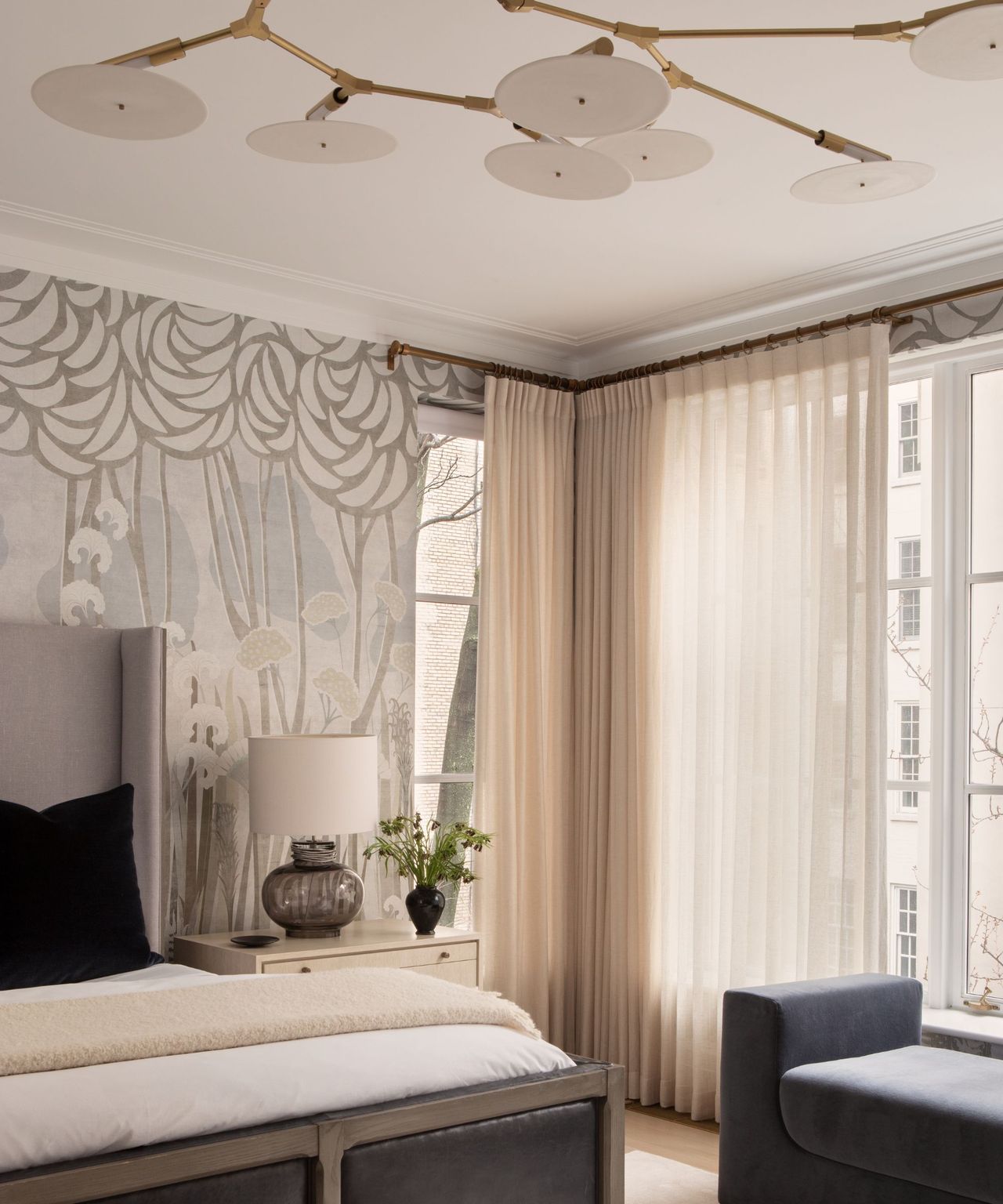 A light and airy bedroom with a gray headboard, blue bench, sheer curtains, expressive wallpaper, and a sculptural light fixture