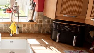 An air fryer sits under kitchen cabinets on a wooden worktop with a sink to the side of it