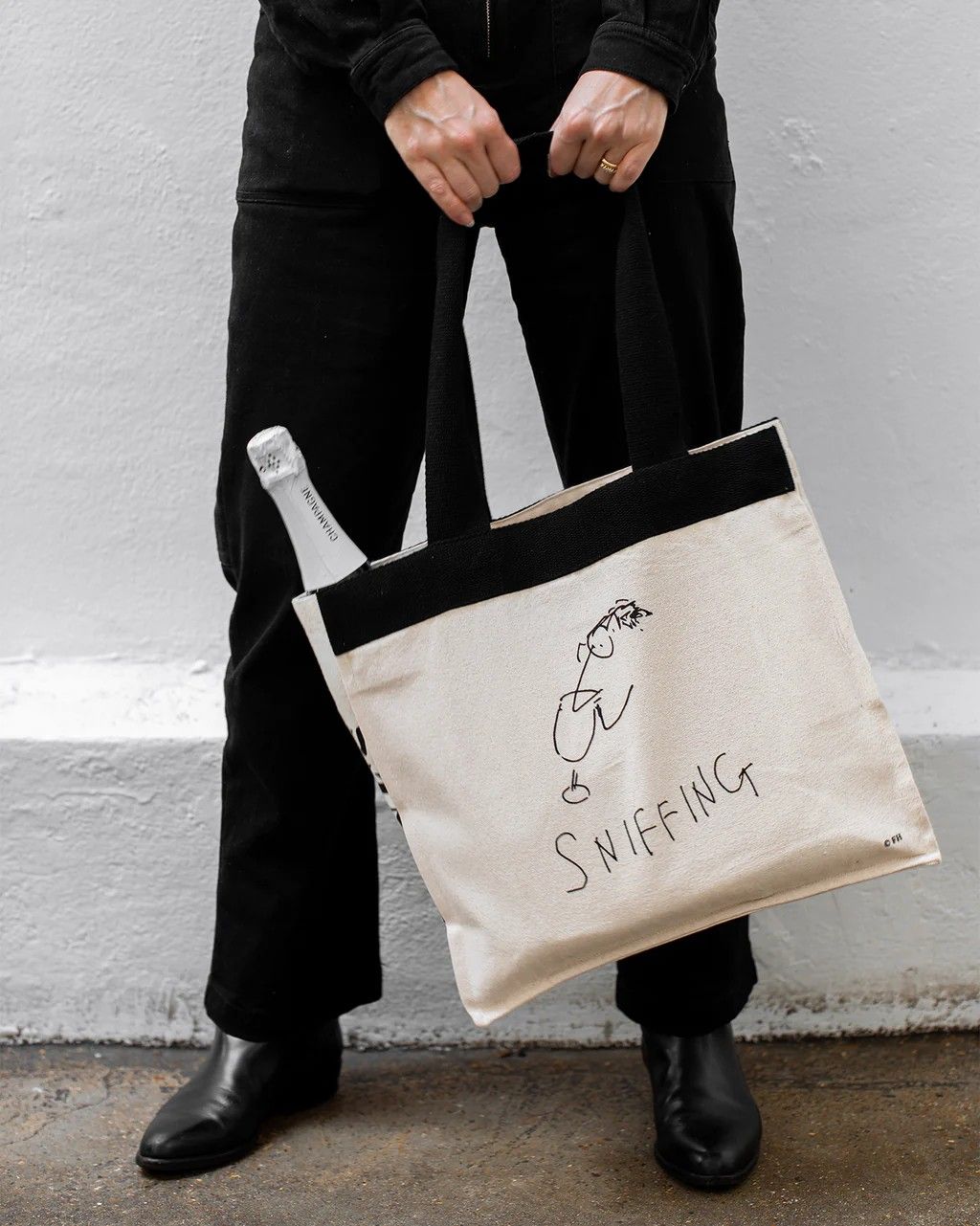 A black and white tote bag with the sketch of a person sniffing wine and the writing "SNIFFING" is being hold by a person entirely dressed in black in an industrial, naturally lit space, and is seen resting while filled with bread and wine on a wooden table in the image next to the previous.