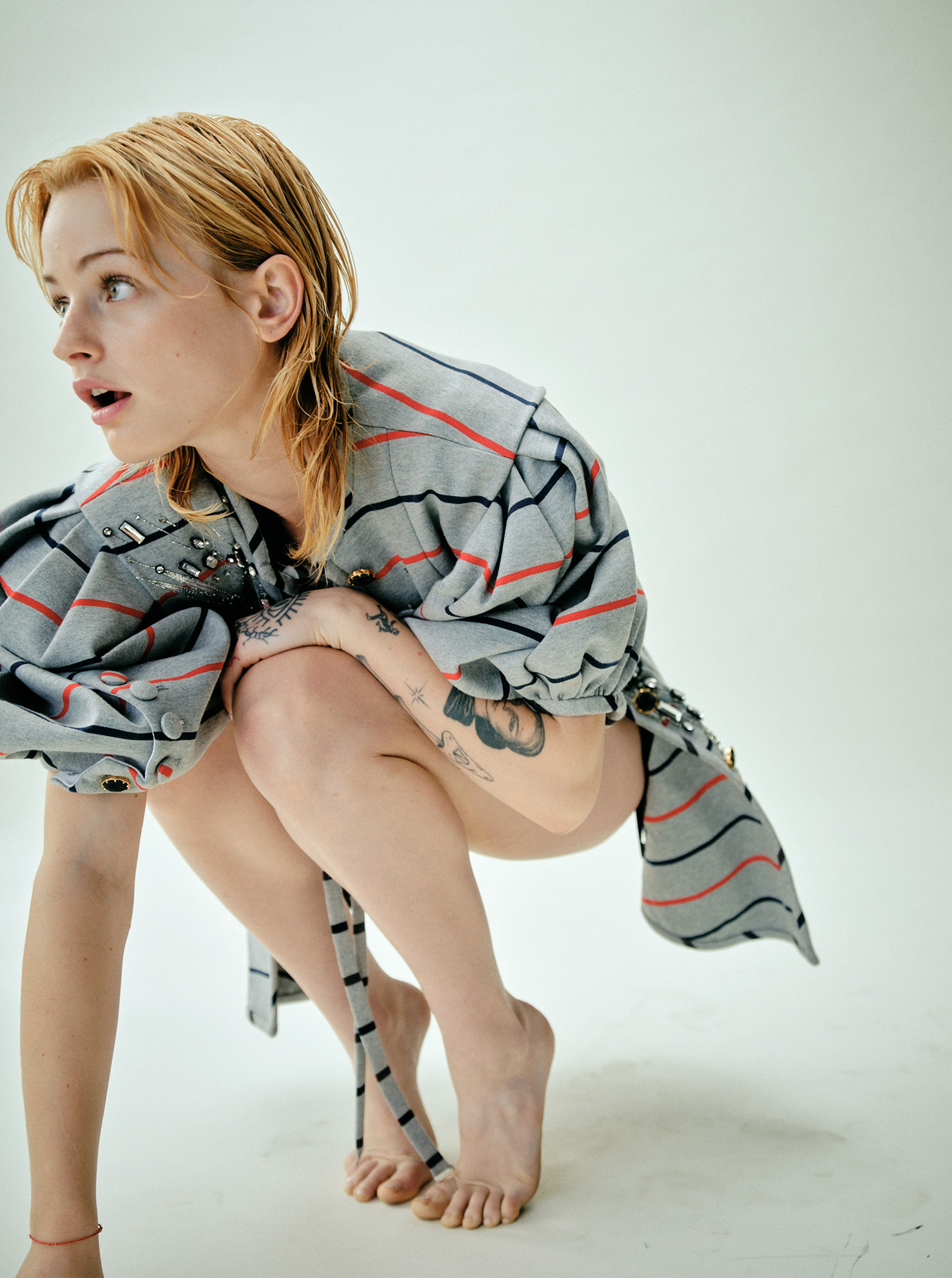 Esther McGregor crouches down balancing on her toes. She is wearing a gray Louis Vuitton overcoat with thin red and navy stripes.