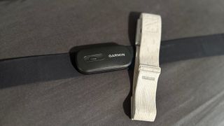 A Garmin HRM 200 chest strap laying on a pillow, with the COROS HRM armband laying perpendicular atop its right side.