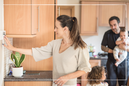 Mother adjusting the thermostat in the kitchen while partner holds child in the background. Partner is also talking to another child standing behind her mother