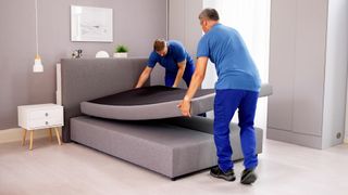 Two men dressed in delivery clothing install a new mattress in a bedroom, placing it on a grey bed frame