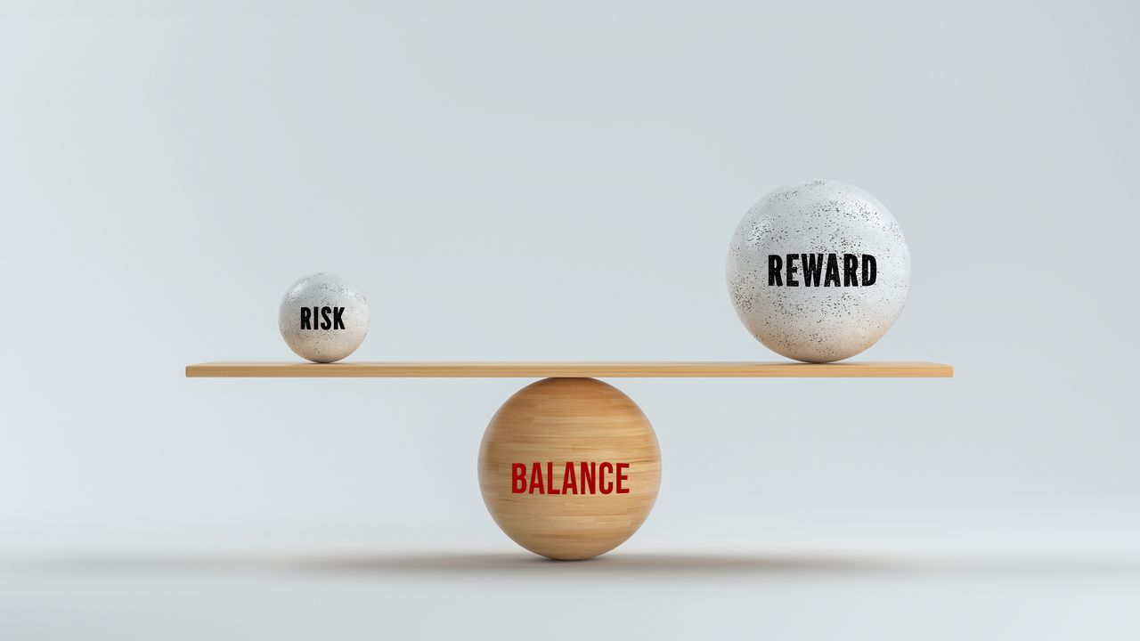 Balls with the words &quot;risk&quot; and &quot;reward&quot; sit on opposite ends of a seesaw sitting on top of a ball that says &quot;balance.&quot;