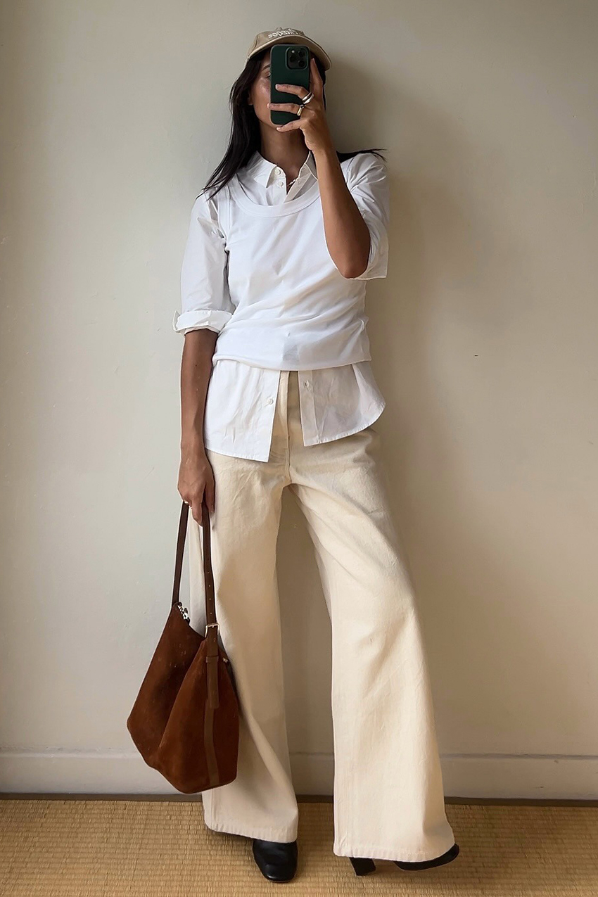 a woman wearing a baseball cap, a button-down, a tank top layered over it, white pants, and black boots