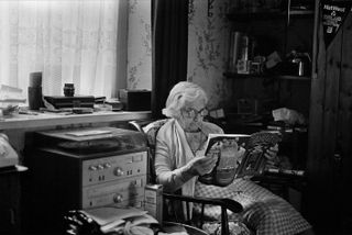 Black and white image of Grandma Thrasher, Swansea, 1984 sat in a wooden arm chair, sitting room, window with net curtains to the left, white window sill with items on top, patterned wall paper, record player under the window