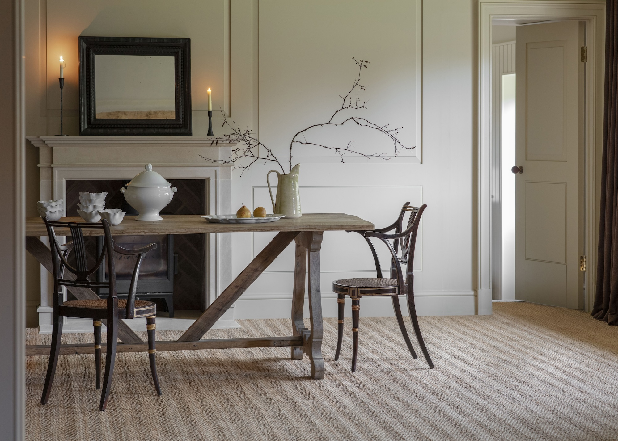 A neutral-toned dining room with a rectangular wooden table and two chairs in front of a fireplace with a mirror above and two lit taper candles with textured carpet flooring