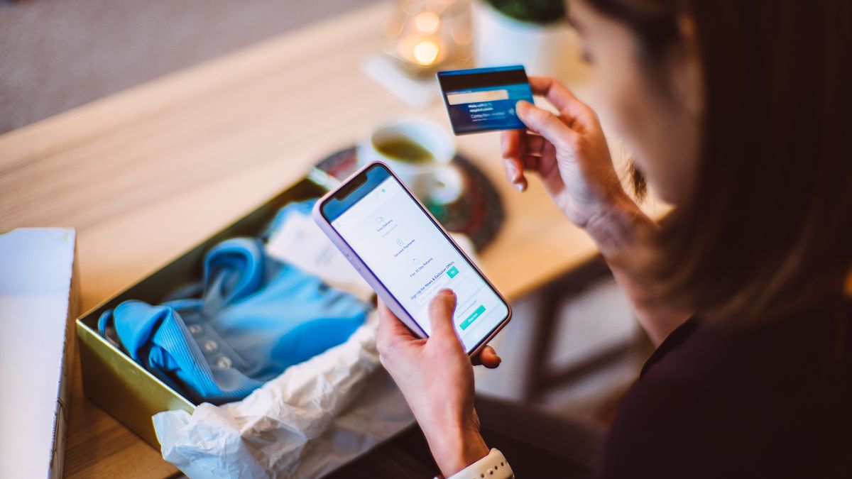 A woman with an open clothing package is holding a smartphone in her left hand and a credit or debit card in her right hand while looking a the smartphone screen.