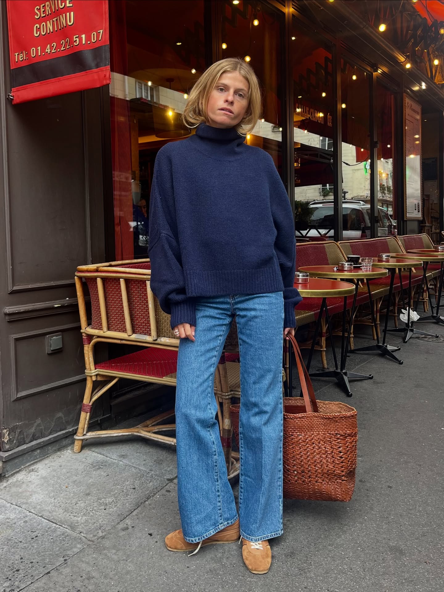 An image of an influencer wearing a navy turtleneck jumper, mid-wash blue jeans and brown suede trainers holding a woven tote in Paris