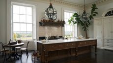 A wooden kitchen island with marble countertops in a kitchen with hanging pendants above. There is also a dining table with chairs on one side of the room