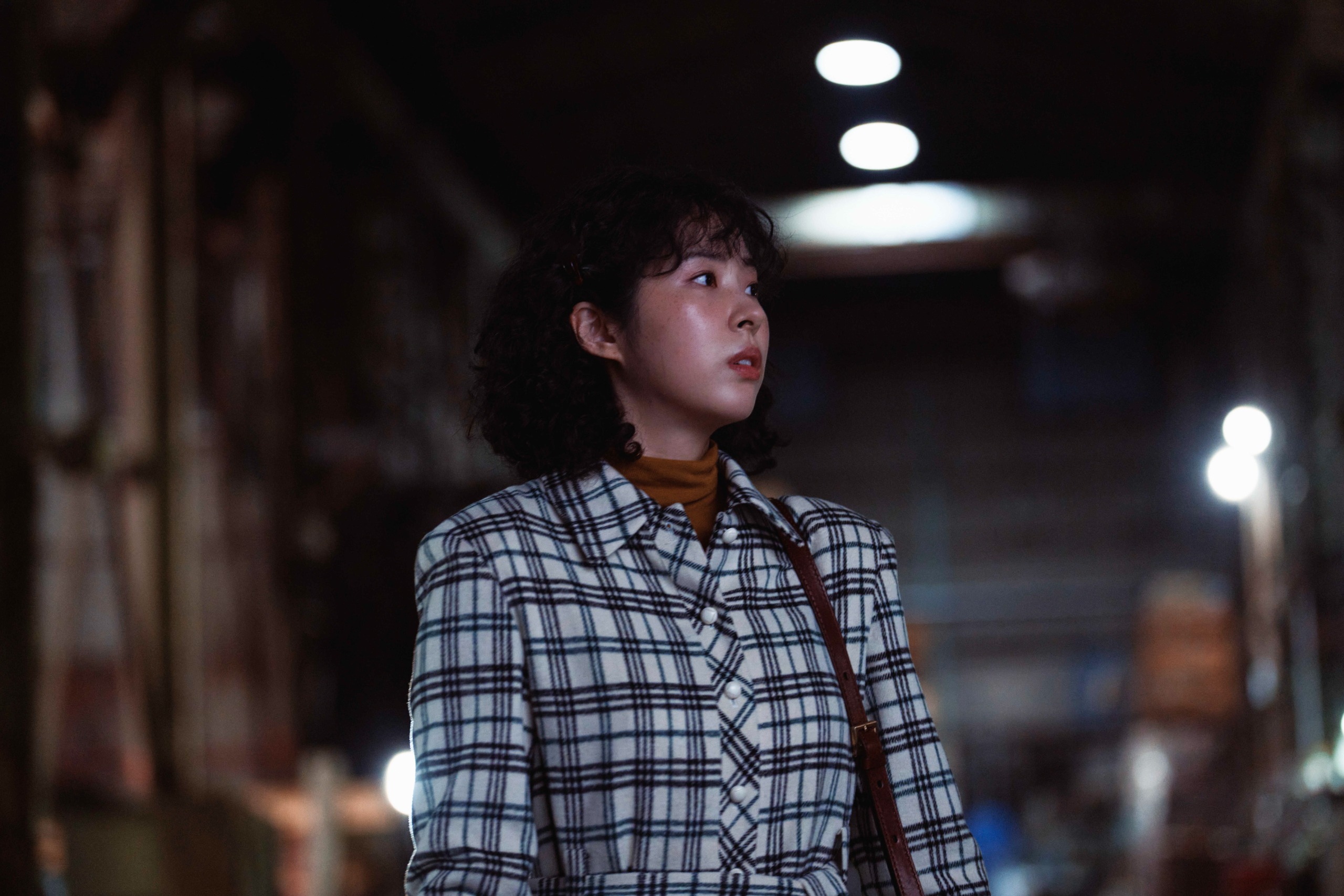 A woman wearing a vintage plaid suit (Seo Eun-soo as Oh Yejin) stands in a large warehouse, in Hulu's 'Made In Korea.'