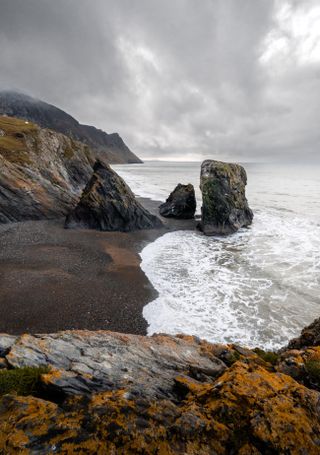 Photo of sea stacks on a gray day. The sea froth is white from slamming against the shore.