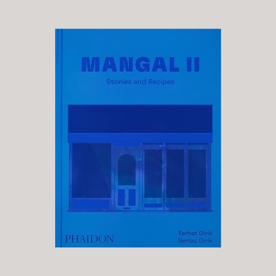 A blue-covered book with the title "MANGAL II: Stories and Recipes" and a sketch of a shop front sits against a pale grey background.