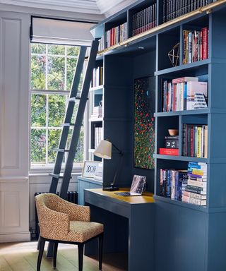 Home office with built in desk and shelving in blue with library ladder and upholstered chair