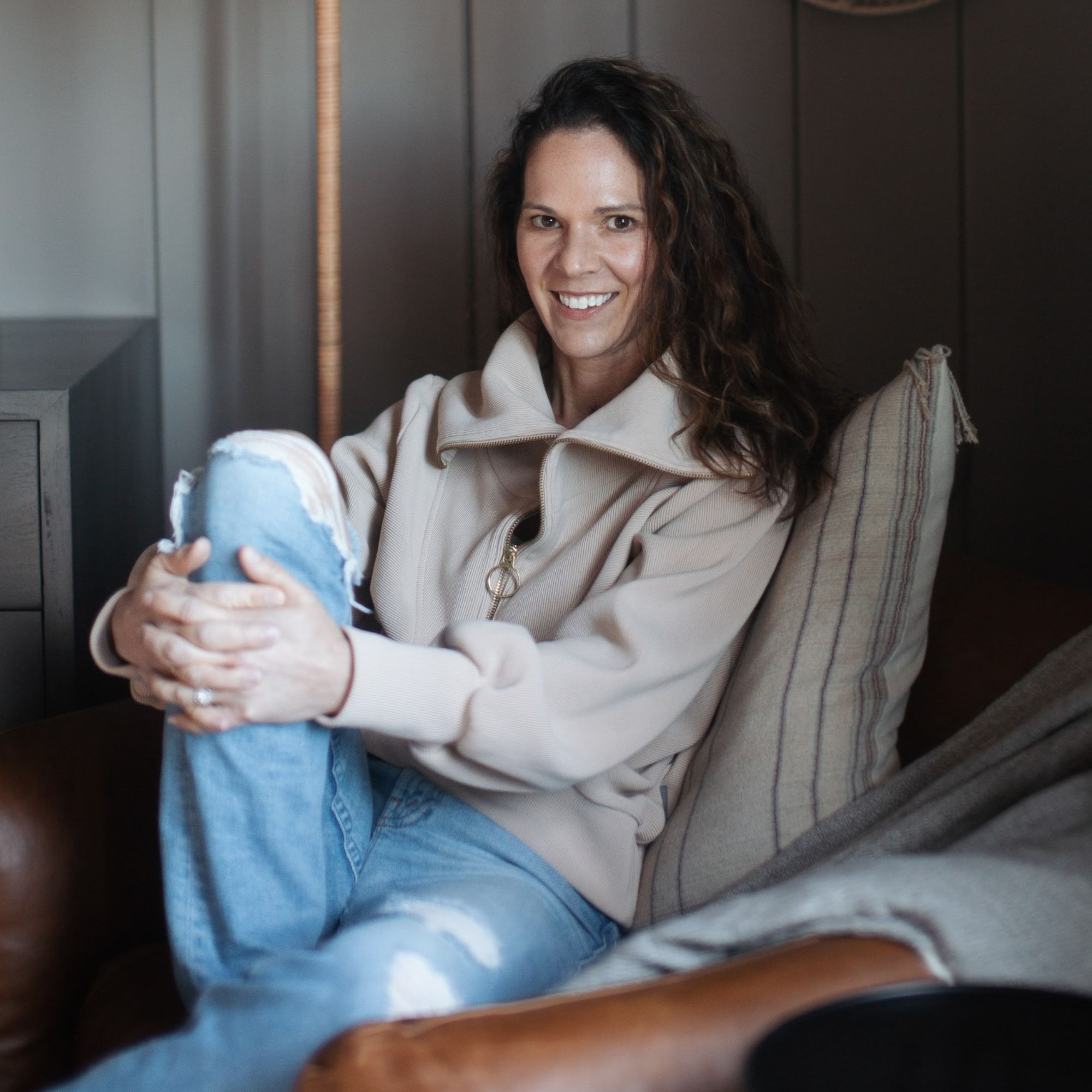 A picture of Amanda Wyatt, a woman with brown hair wearing a cream jacket with light blue ripped jeans on, sitting on a light brown leather armchair with a large gray throw pillow behind her