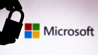 The Microsoft logo against a white background out of focus, with the silhouette of a hand holding a padlock to the left of frame in focus.