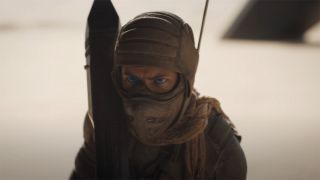 Still from the movie Dune: Part 2. A Fremen person with bright blue eyes is completely covered in armor. They are holding what looks like a big weapon, resting it on their shoulder.