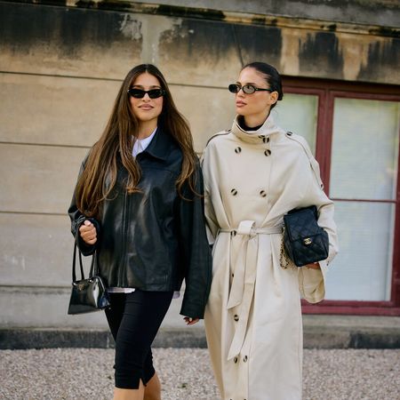 Two women walk and pose. One wears black coat and the other wears a beige trench. They both carry black handbags. 