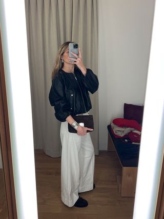 Eliza Huber in the dressing room at Madewell's new SoHo flagship in New York wearing a cropped leather balloon-sleeve jacket with wide-leg white jeans, brown suede loafers, and a brown suede bag.