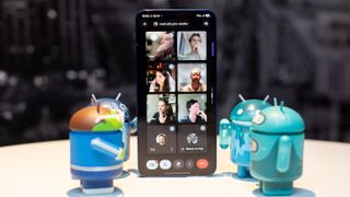 Samsung Galaxy S25 Plus in Navy Blue showing a TechRadar Google Meet video call, standing in between four adorable Android figurines