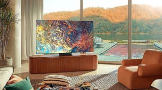 The Samsung QN900A TV in a brightly lit room with floor to ceiling windows behind it and a view of a forest
