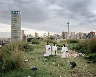 Three people in white robes kneeling on grass with scattered rubbish all over under a cloudy sky. There are multiple buildings in the distance