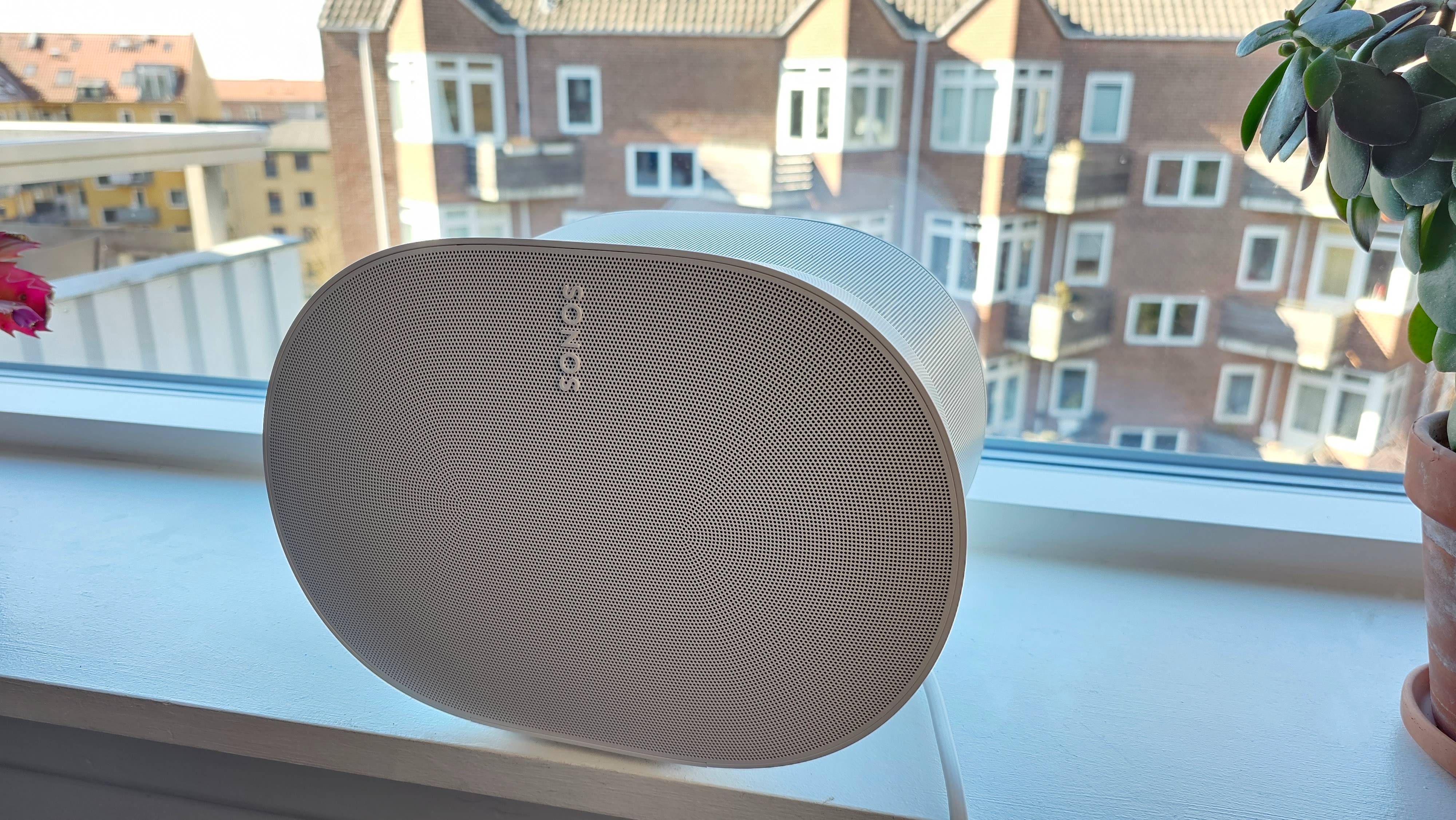En hvid Sonos Era 300 set forfra i en vindueskarm med nabobygning i baggrunden