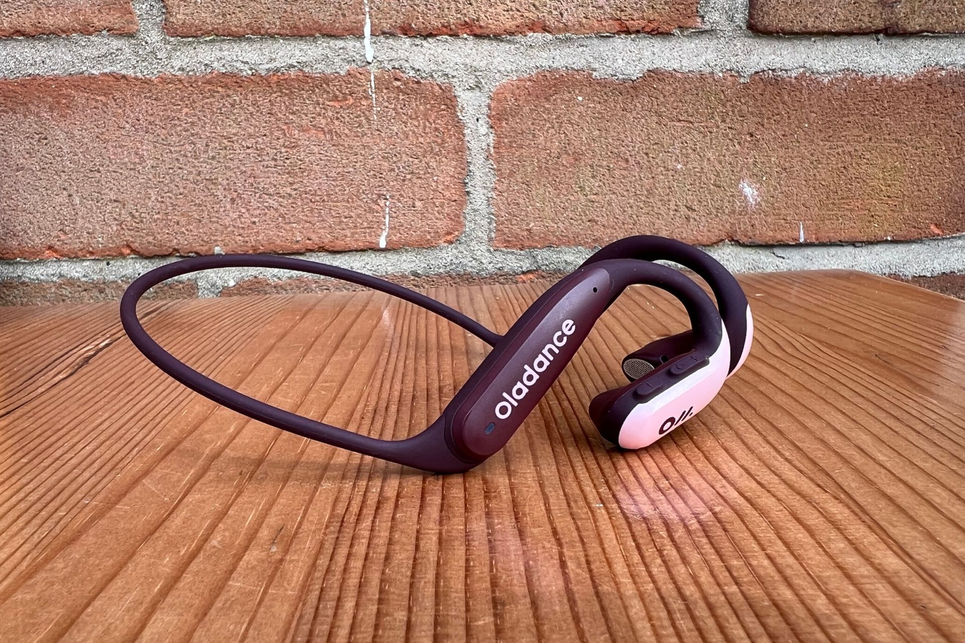 A pink pair of Oladance OWS Sports headphones are placed on a wooden surface with a brick wall behind