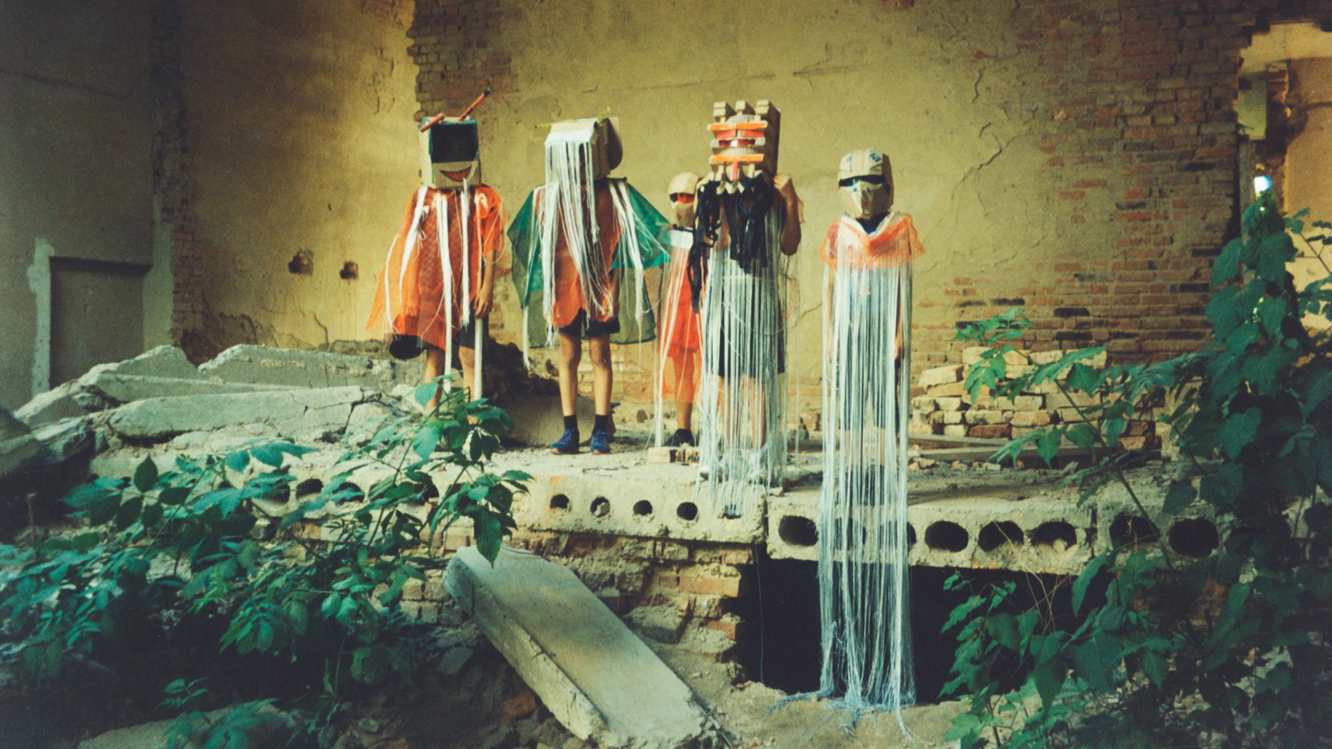 Four children in homemade superhero costumes with box-like masked heads and trailing fringes stand on rubble in a roofless brick ruin overgrown with plants.