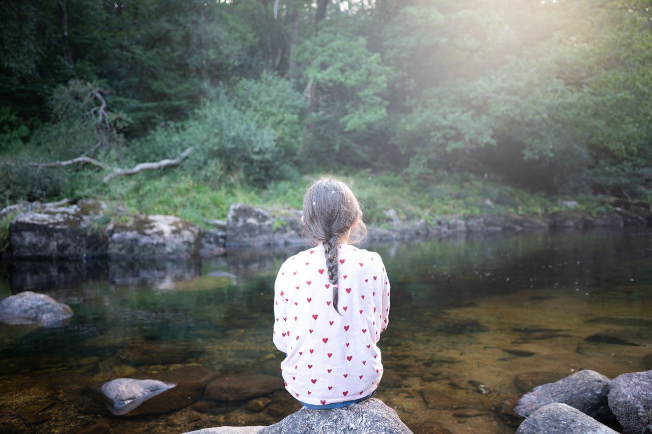 A girl sits on a rock alongside a river, there's lens flar making a soft haze effect