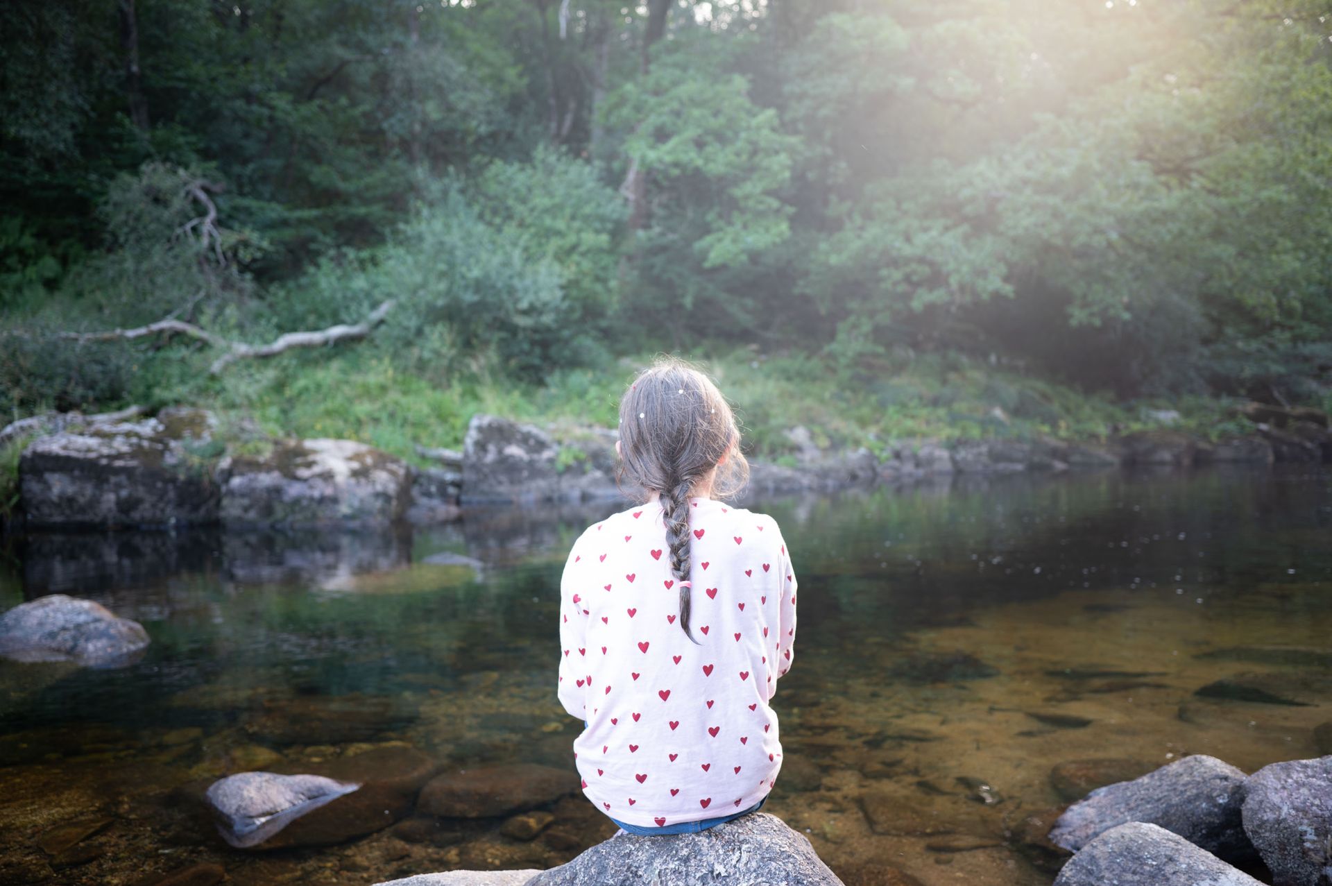 A girl sits on a rock alongside a river, there's lens flar making a soft haze effect