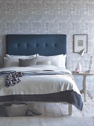 gray and blue bedroom with gray linear wallpaper and blue bed, white artwork, white bedding, gray accessories, side table, chrome desk lamp, basket