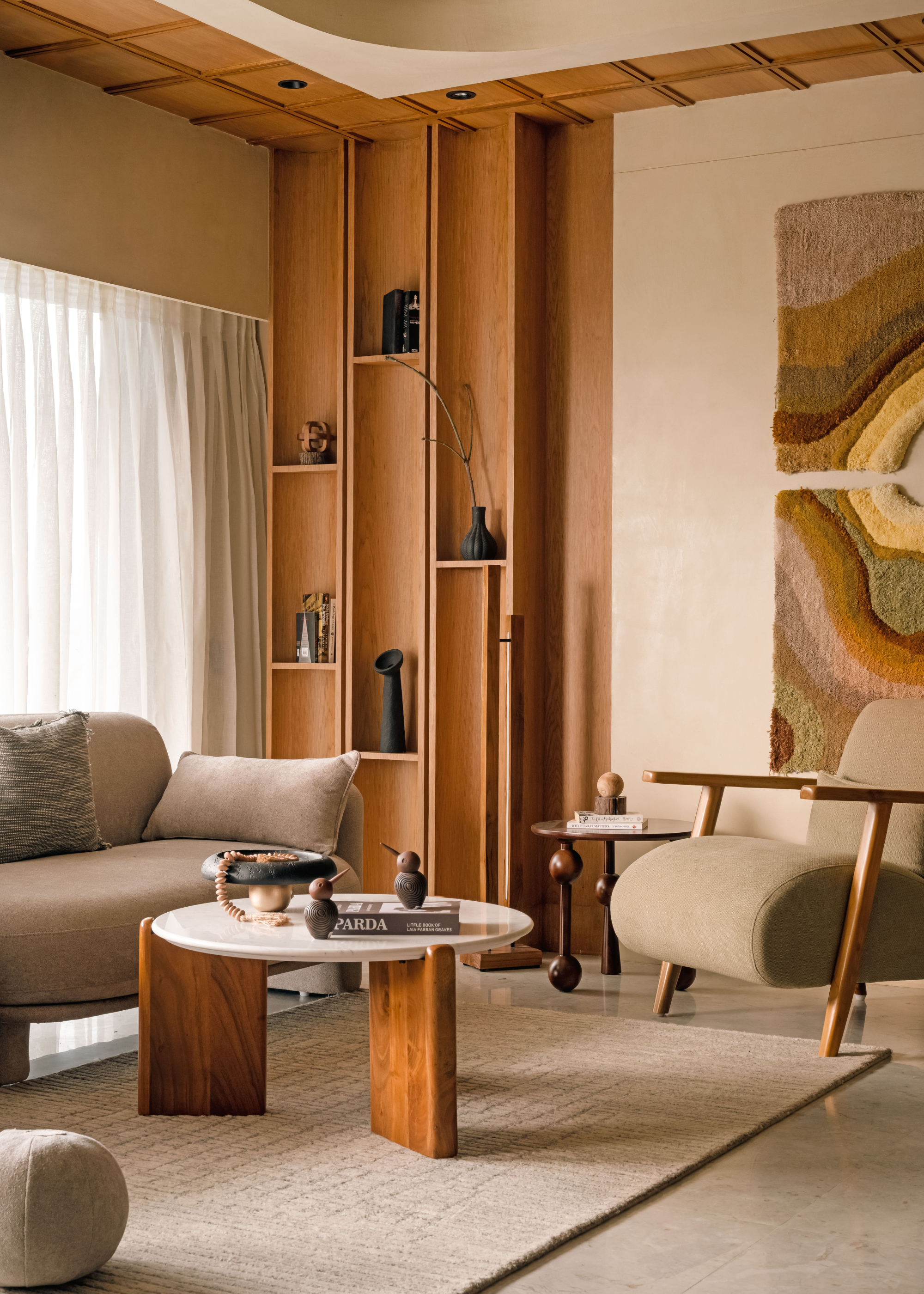 A bright earthy living room with a wooden shelving system, a hanging textured tapestry, a beige and wood armchair, a beige sofa, a round coffee table on a gray rug